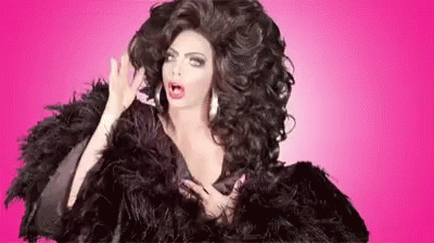 A Drag Queen With Very Long Hair Is Wearing A Black Fur Coat And Feathers . GIF