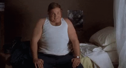 A Fat Man Is Sitting On A Bed In A Bedroom Wearing A White Tank Top . GIF