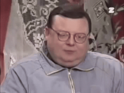 A Fat Man Wearing Glasses And A Blue Jacket Is Sitting In Front Of A Window . GIF