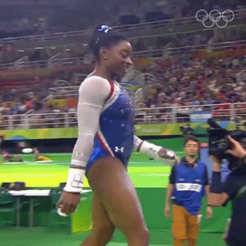 A Female Gymnast Is Walking In Front Of A Crowd While Wearing A Blue And White Leotard . GIF
