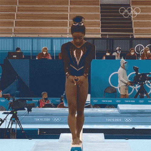 A Female Gymnast Stands On A Balance Beam In Front Of A Tokyo 2020 Sign GIF