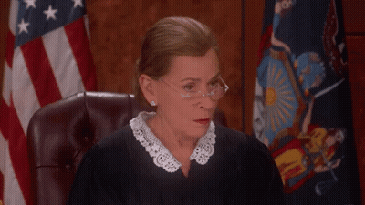A Female Judge Wearing Glasses And A Lace Collar Sits In Front Of An American Flag GIF