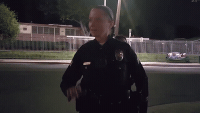 A Female Police Officer Stands In Front Of A Fence And A Building At Night GIF