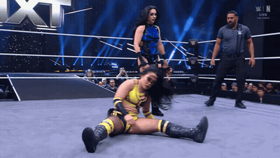 A Female Wrestler Is Laying On The Ground In A Wrestling Ring While Another Wrestler Stands Behind Her GIF