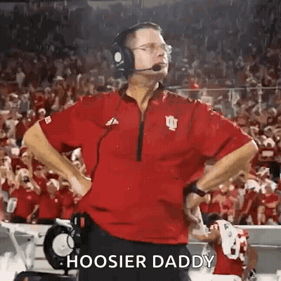 A Football Coach Wearing Headphones And A Red Shirt Is Standing On The Sidelines Of A Field . GIF