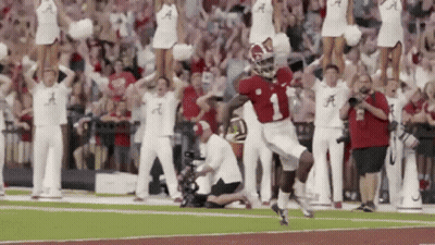 A Football Player Is Running On The Field With Cheerleaders Behind Him And A Crowd Watching . GIF