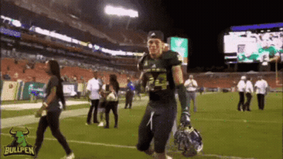 A Football Player Is Walking On A Field With A Helmet In His Hand . GIF