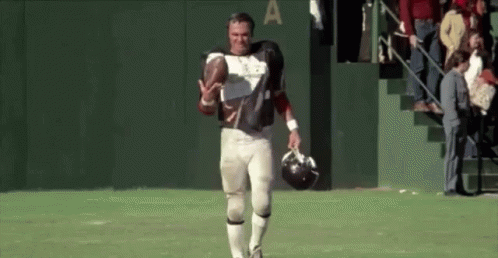 A Football Player Is Walking On The Field Holding A Helmet And A Football . GIF