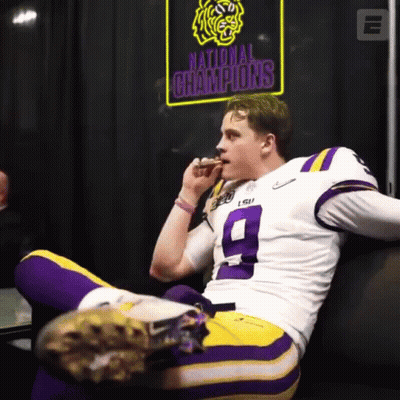 A Football Player Sitting On A Couch With A Sign That Says National Champions Behind Him GIF