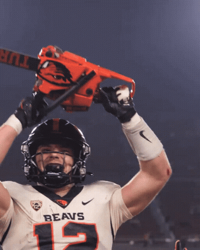 A Football Player With The Number 12 On His Jersey Is Holding A Chainsaw Over His Head GIF
