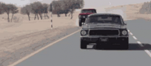 A Ford Mustang Is Driving Down A Desert Road Next To A Red Truck GIF