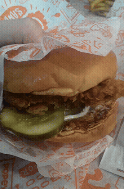 A Fried Chicken Sandwich With A Pickle On Top Is Wrapped In Paper That Says Shakes GIF