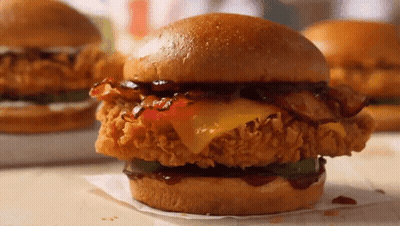 A Fried Chicken Sandwich With Cheese Bacon And Pickles On A Bun GIF