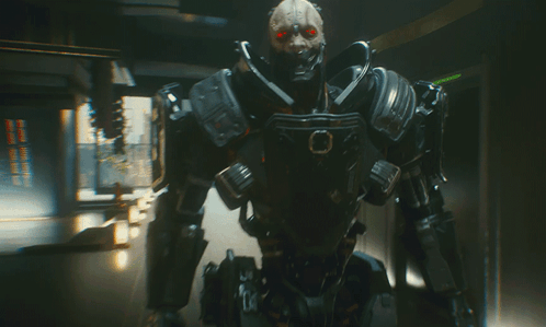 A Futuristic Robot With Red Eyes Is Standing In A Dark Room GIF