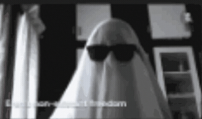 A Ghost Wearing Sunglasses And A Sheet Is Standing In A Room . GIF