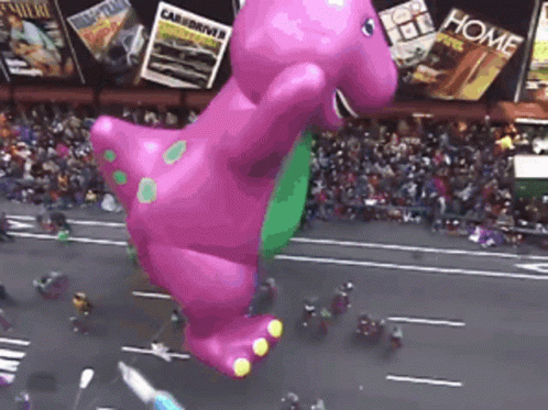 A Giant Balloon Of Barney The Dinosaur Is Flying Over A Street GIF