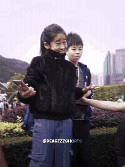 A Girl In A Black Sweater Is Holding A Cell Phone While Standing Next To A Boy In A Blue Jacket GIF