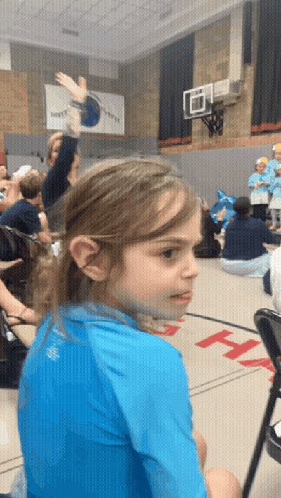 A Girl In A Blue Shirt Is Sitting In A Gym With A Basketball Hoop On The Wall GIF