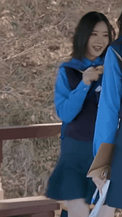 A Girl In A Blue Vest And Skirt Is Holding A Clipboard And Smiling While Standing Next To Another Girl . GIF