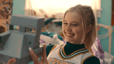 A Girl In A Cheerleader Uniform Is Holding A Polaroid Camera In Her Hand GIF