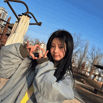 A Girl In A Grey Jacket Is Making A Heart With Her Hands GIF