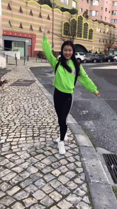 A Girl In A Neon Green Sweatshirt And Black Pants Is Standing On A Cobblestone Sidewalk . GIF