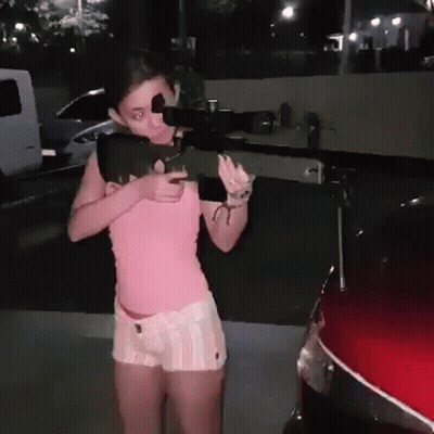 A Girl In A Pink Shirt And White Shorts Is Holding A Gun In Front Of A Red Car . GIF