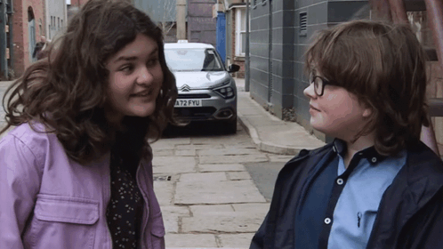 A Girl In A Purple Jacket Stands Next To A Boy In A Blue Shirt With A License Plate That Says A72fyu GIF
