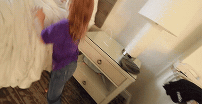 A Girl In A Purple Shirt Is Standing Next To A Nightstand With A Lamp On It GIF
