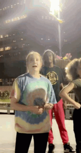 A Girl In A Tie Dye Shirt Stands In Front Of A Group Of People Dancing GIF
