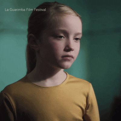 A Girl In A Yellow Shirt Is Standing In Front Of A Green Wall And The Words La Guarimba Film Festival Are Above Her GIF