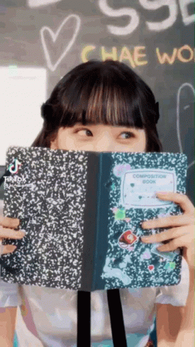 A Girl Is Covering Her Face With A Composition Book GIF