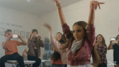 A Girl Is Dancing In Front Of A Group Of People And The Word Brat Is On The Bottom Left GIF