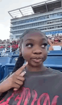 A Girl Is Pointing At Her Face While Sitting In The Stands At A Baseball Game . GIF