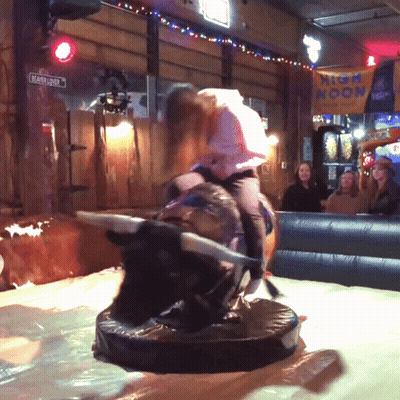 A Girl Is Riding A Bull In A Room With A Sign That Says High Noon On It GIF