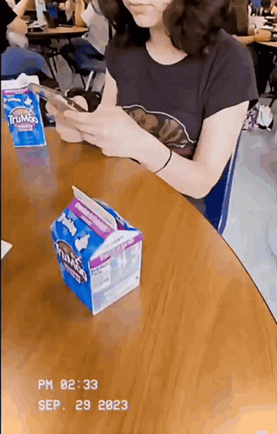 A Girl Is Sitting At A Table With A Carton Of Trumps Milk GIF