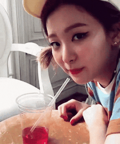 A Girl Is Sitting At A Table With A Cup Of Liquid And A Straw GIF