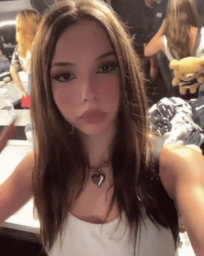 A Girl Is Taking A Selfie With A Teddy Bear In The Background While Wearing A Heart Necklace . GIF