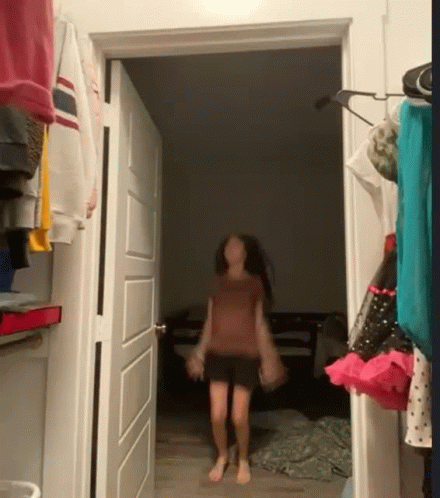 A Girl Is Walking Through A Doorway In A Room With Clothes Hanging On The Wall . GIF