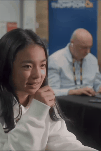 A Girl Sits At A Table With A Man Behind Her And A Sign That Says Troop Room On It GIF