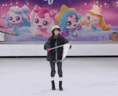 A Girl Stands On A Ice Rink Holding A Flag In Front Of A Wall With Cartoon Characters On It GIF