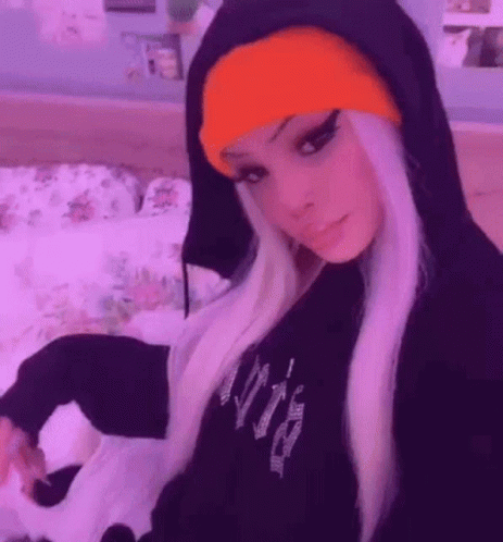 A Girl Wearing A Black Hoodie And An Orange Beanie Is Sitting On A Bed . GIF