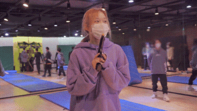A Girl Wearing A Mask And A Purple Sweatshirt Is Holding A Gun In A Gym GIF