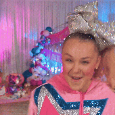 A Girl Wearing A Pink Hoodie And A Large Bow In Her Hair Is Standing In A Room . GIF