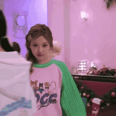 A Girl Wearing A Pink Shirt And A Green Sweater Is Standing In A Pink Room . GIF
