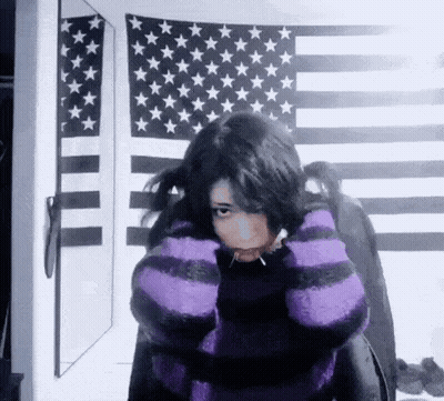 A Girl Wearing A Purple And Black Striped Sweater Is Standing In Front Of An American Flag And A Mirror . GIF