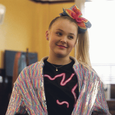 A Girl Wearing A Rainbow Jacket And A Bow In Her Hair Smiles GIF