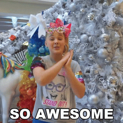 A Girl Wearing A T-Shirt That Says " Stand Out " Is Clapping In Front Of A Christmas Tree GIF