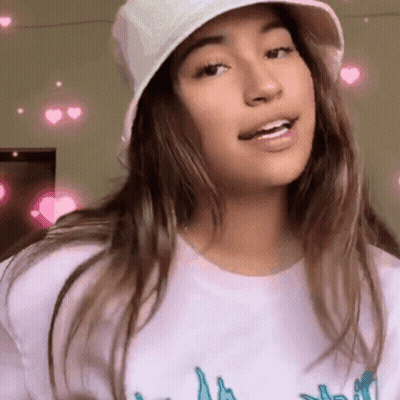 A Girl Wearing A White Hat And A White T-Shirt Is Smiling And Looking At The Camera . GIF