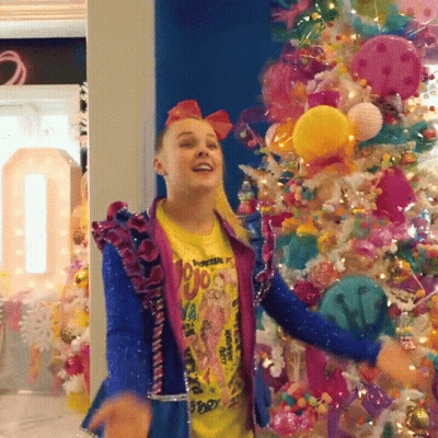 A Girl Wearing A Yellow Shirt That Says ' Jojo ' On It Is Dancing In Front Of A Christmas Tree GIF